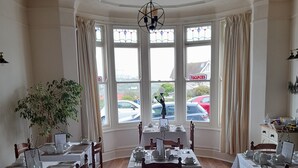 A dining area with tables set for service, large windows with curtains, and a view of parked cars outside.