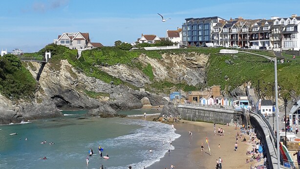 A coastal beach scene with a rocky cliff, a promenade, and buildings in the background.