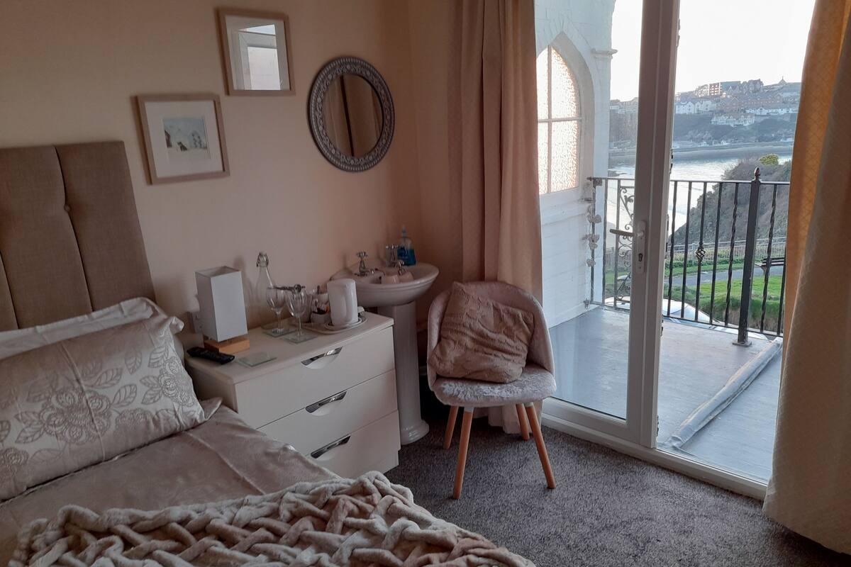 A bedroom with a bed, a bedside table with a lamp, a chair, and a view of the outside.