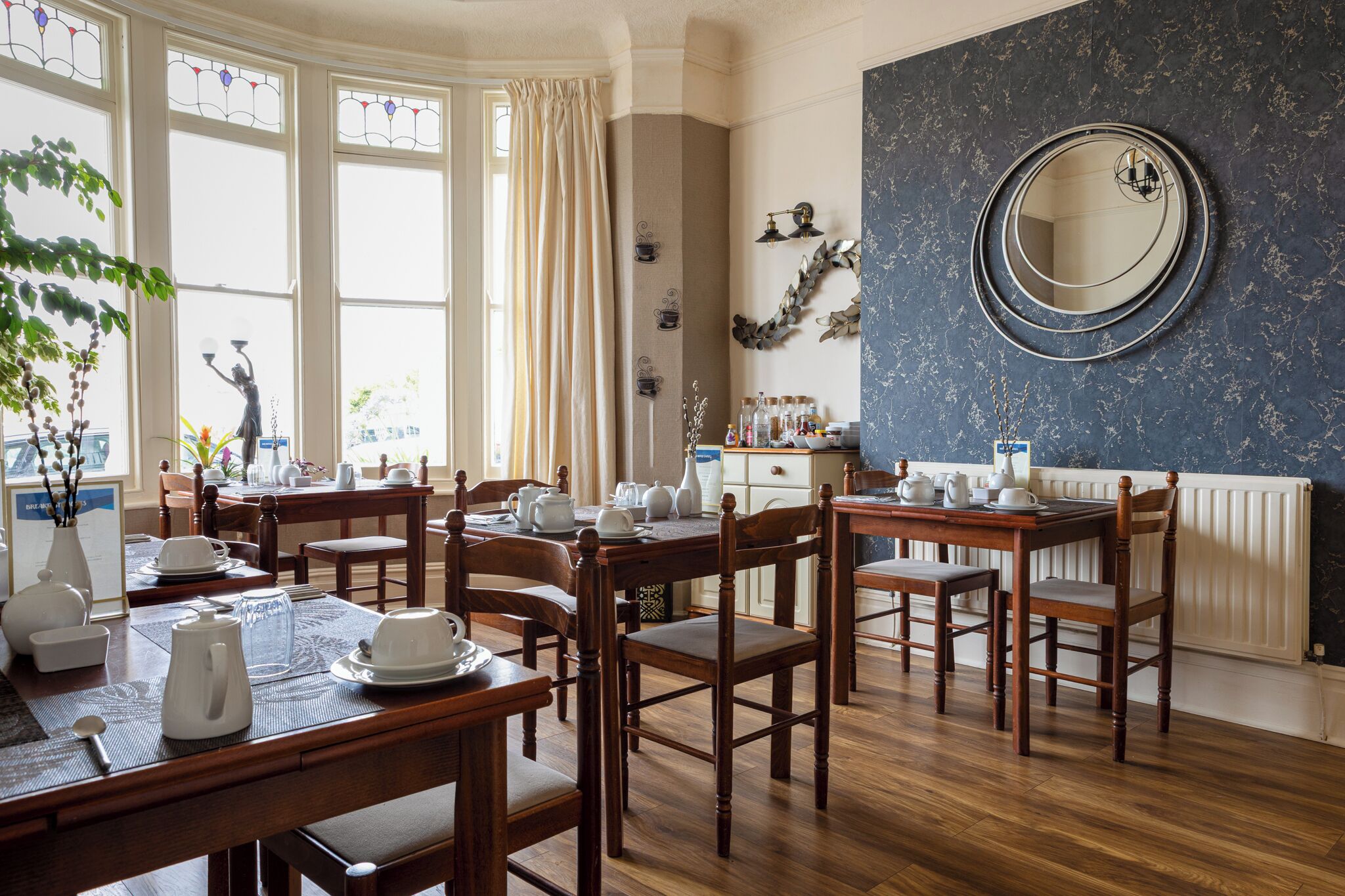 A dining area with wooden tables and chairs, a large mirror on the wall, and a window with curtains.