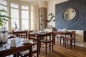 A dining area with wooden tables and chairs, a large mirror on the wall, and a window with curtains. - Tregella Guest House (Newquay)