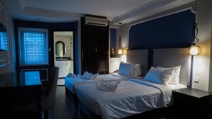 Habitación de hotel con una cama grande, un escritorio y una silla. Hay una ventana con persianas, un espejo y una mesita de noche con lámpara.
