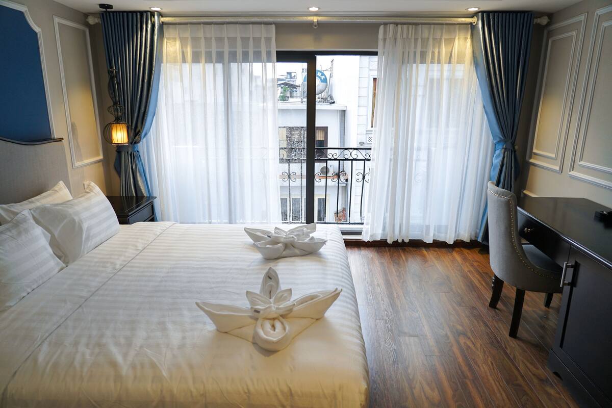 Une chambre d’hôtel avec un grand lit, un bureau et une vue sur la ville à travers de fines rideaux.