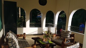Un área de estar con muebles de madera y cojines estampados, una mesa con una planta y ventanas arqueadas con vistas a follaje.