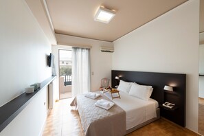 A modern hotel room with a large bed, a bedside table with a lamp, a flat-screen TV mounted on the wall, and a balcony with a view.