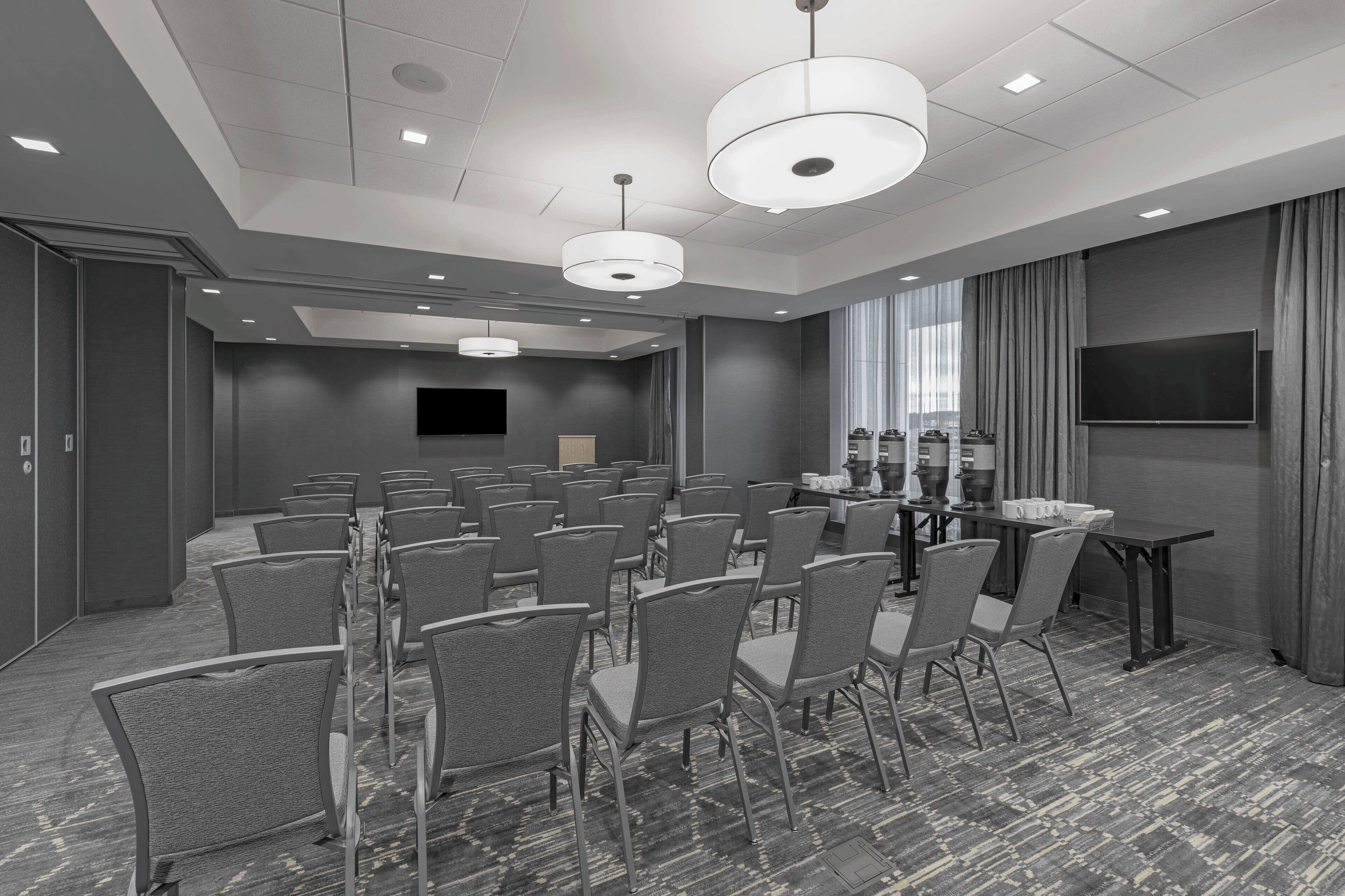 A conference room with rows of chairs, a table, a flat-screen TV, and a large window with curtains.