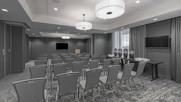 A conference room with rows of chairs, a table, a flat-screen TV, and a large window with curtains.