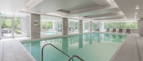 A modern indoor swimming pool with clear glass walls, a tiled floor, and a few lounge chairs around the pool.