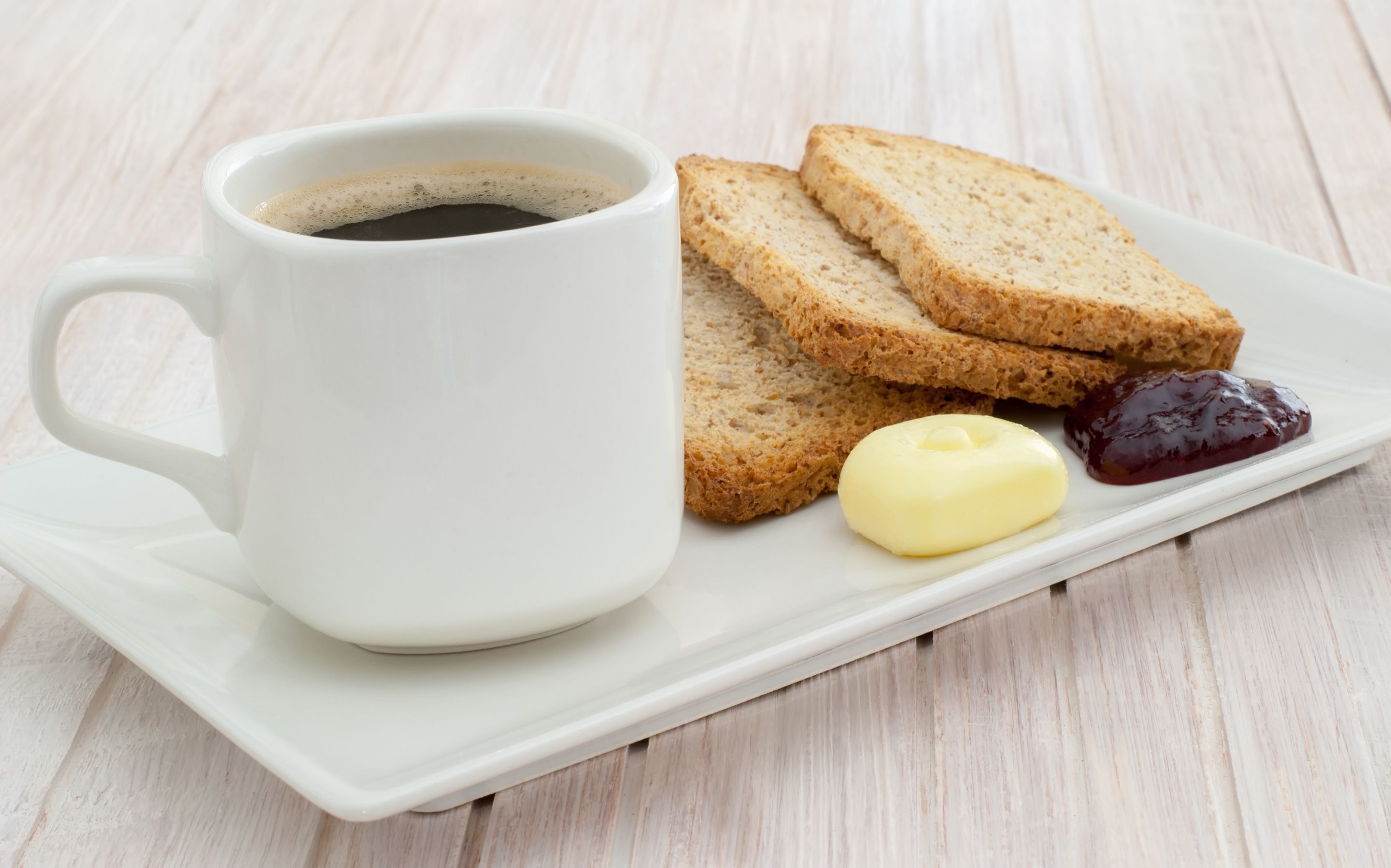 Une tasse en céramique blanche contenant du café, deux tranches de pain complet, une petite portion de beurre et une cuillère de confiture, le tout disposé sur un plateau blanc.