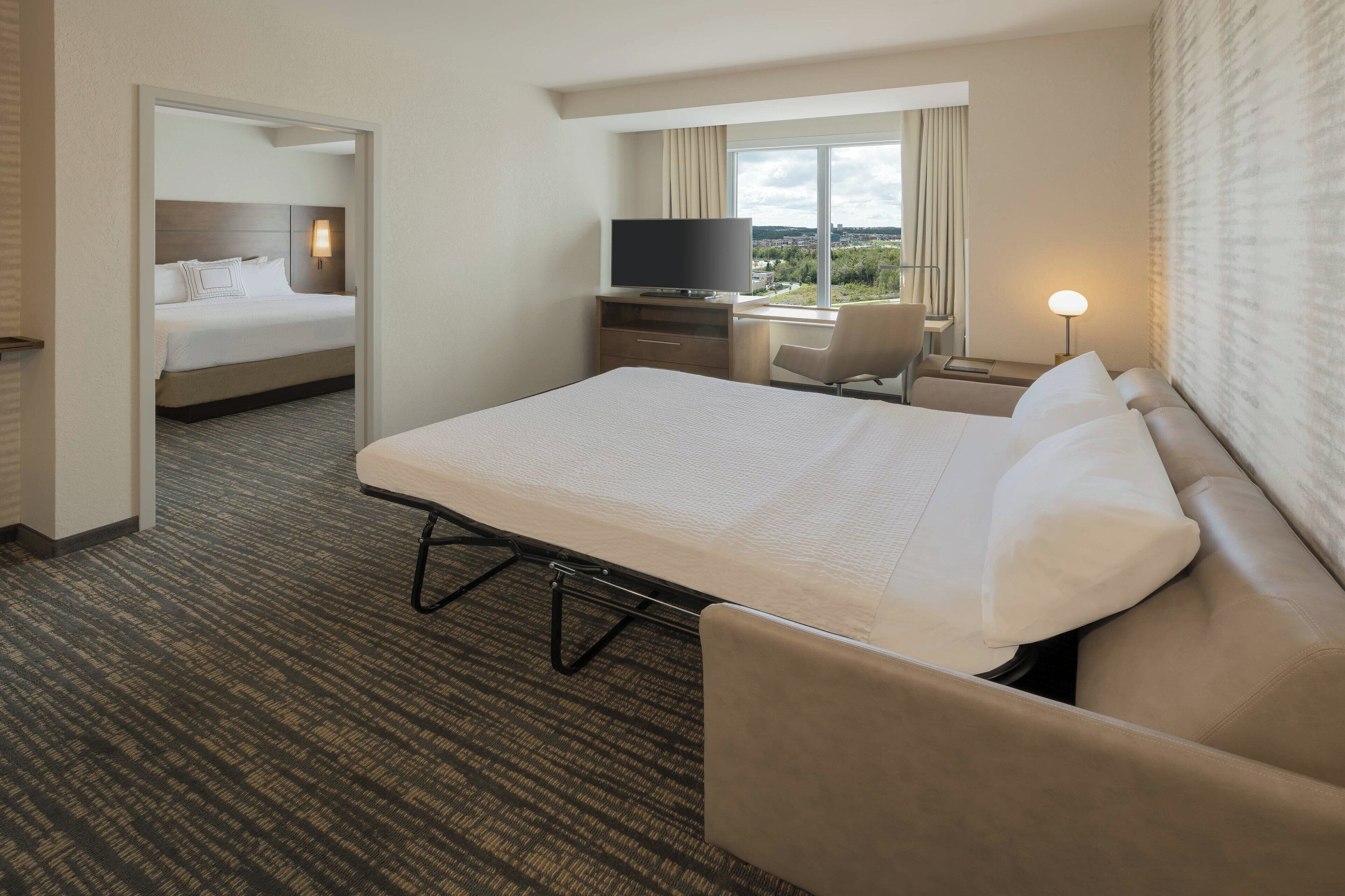 A hotel room with a large bed, a desk with a television, a chair, and a view of the city.