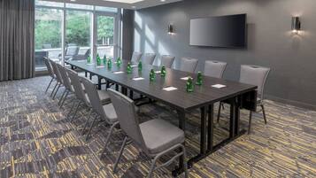 A conference room with a long table, chairs, a flat-screen TV, and a large window with a view of trees.