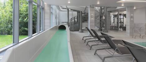 A modern indoor swimming pool with a green lane, large windows, and a staircase leading to a gym area.