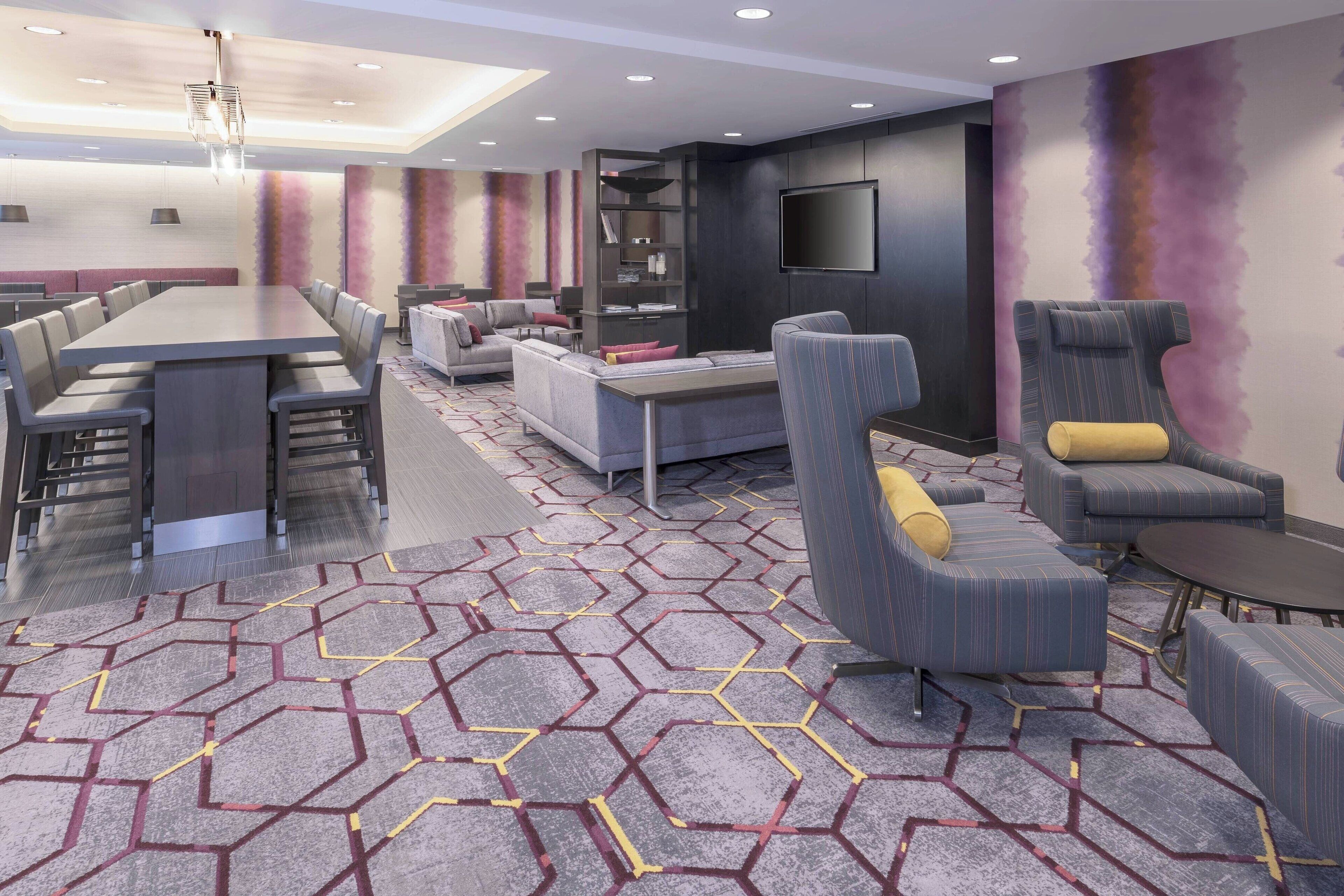 A modern hotel lobby with a long dining table, sofas, a bookshelf, and a flat-screen TV on the wall.
