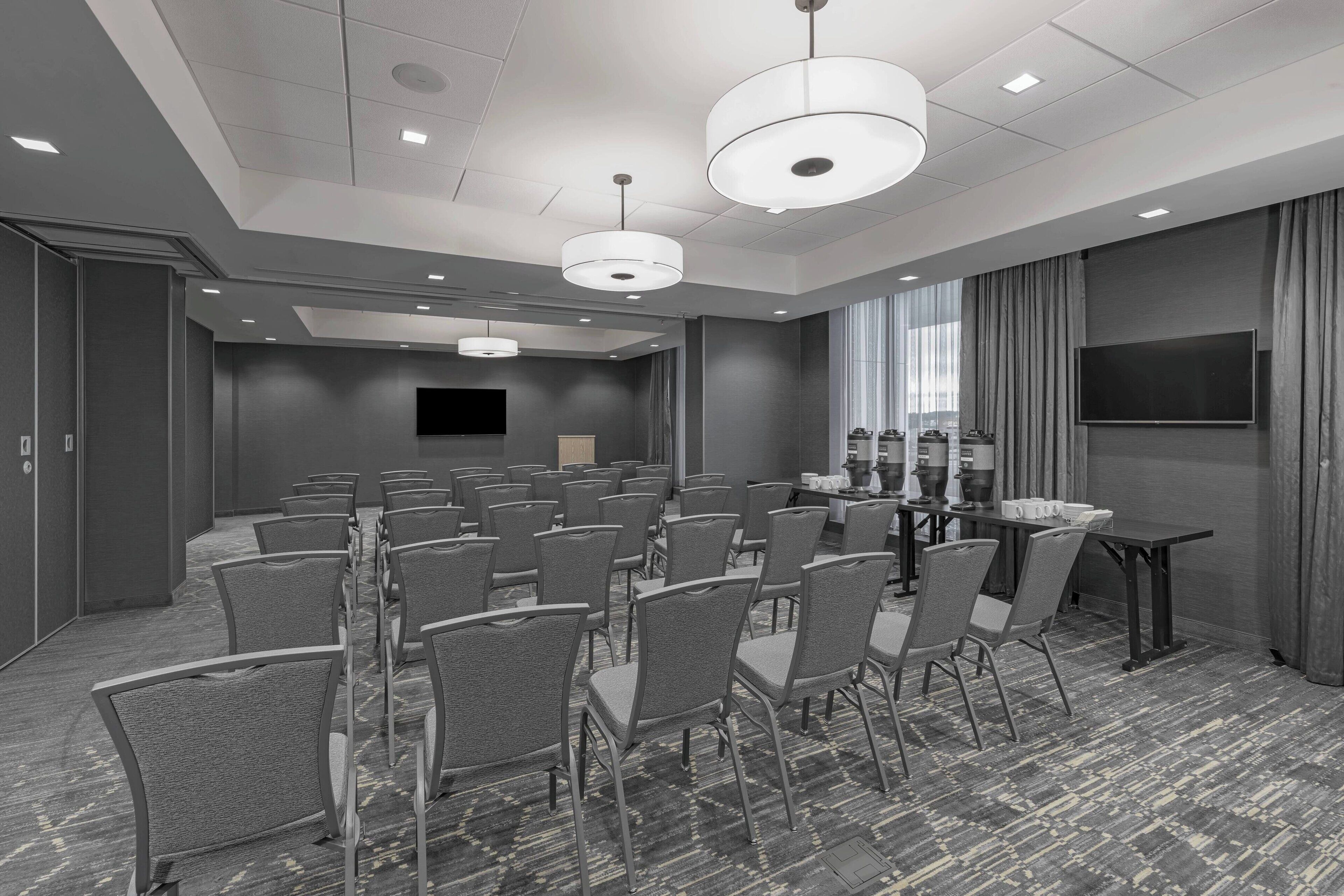 A conference room with rows of chairs, a table with chairs, a flat-screen TV, and a large window with curtains.