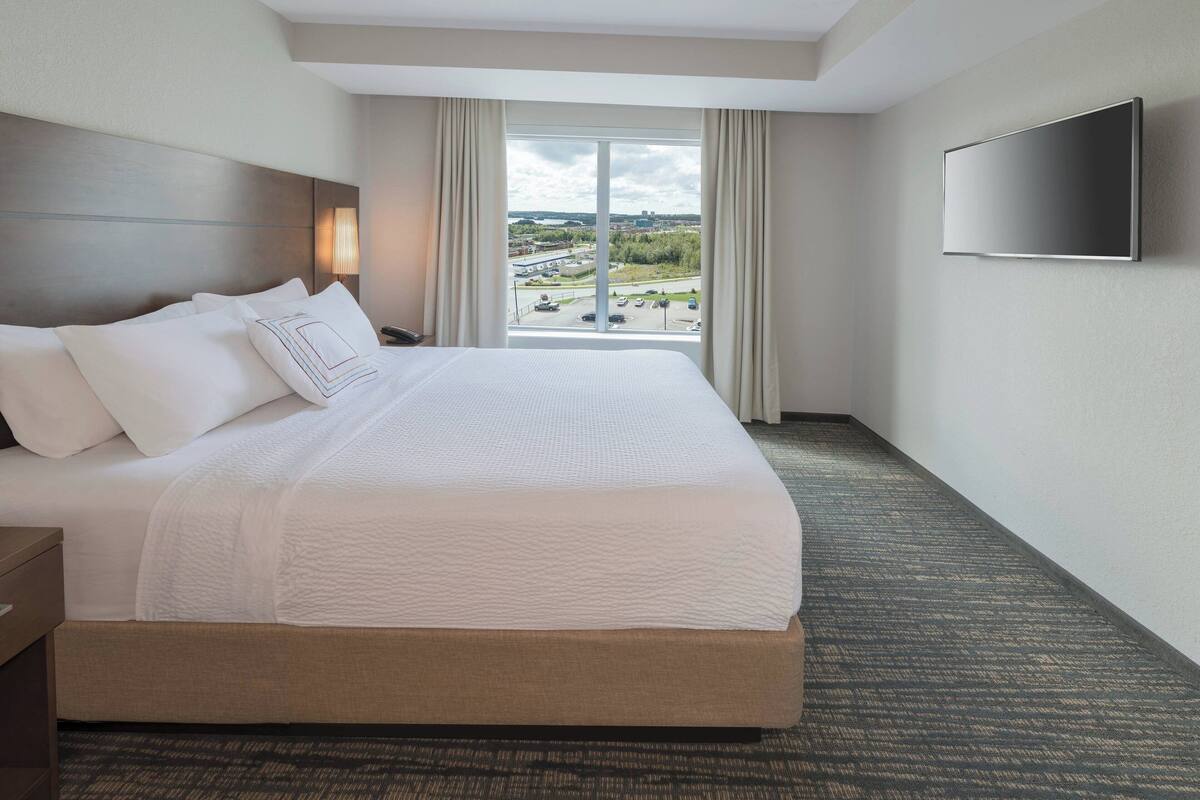 A hotel room with a large bed, a flat-screen TV mounted on the wall, and a view of the outside through a large window.