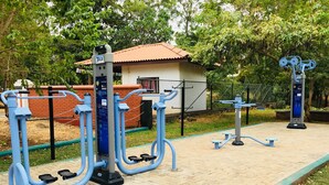 Outdoor exercise equipment in a park with a brick path, a small building, and trees in the background.