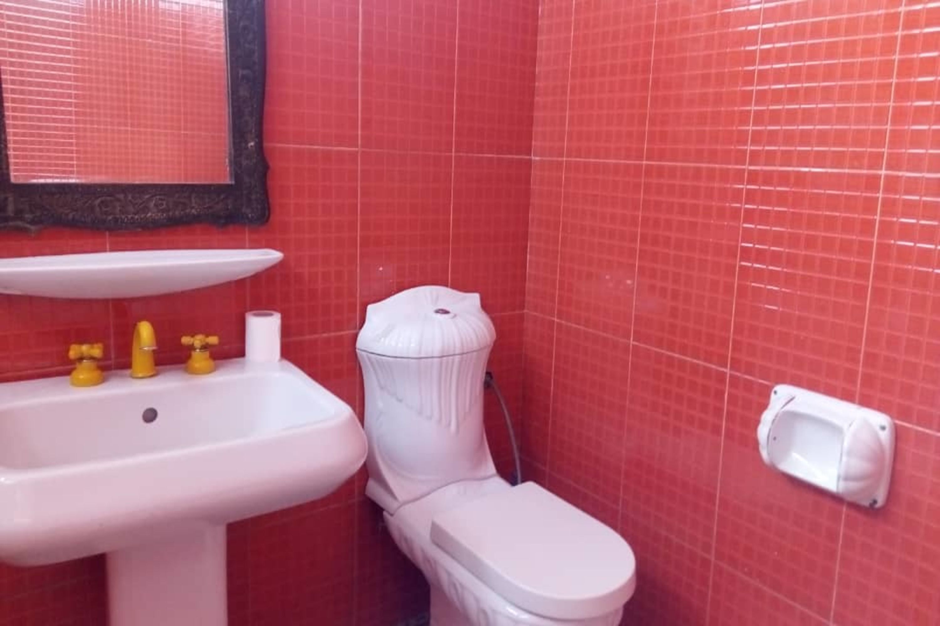 Un baño con un lavamanos blanco, grifos amarillos, un inodoro blanco y paredes de azulejos rojos.