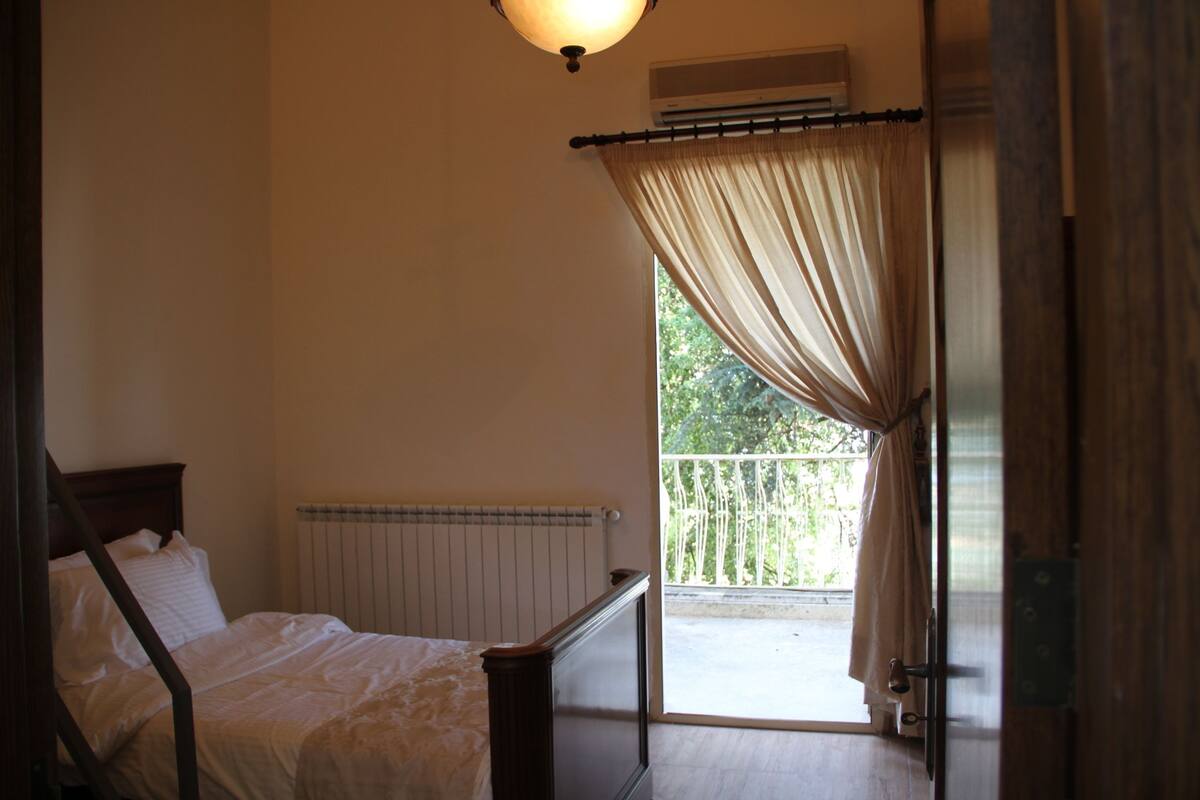 Ein Schlafzimmer mit Bett, Heizkörper, einem Fenster mit Vorhängen und einer offenen Tür, die auf einen Balkon führt.