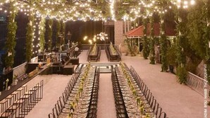 Un espace extérieur aménagé pour les repas, avec des guirlandes lumineuses et de la verdure, des tables dressées pour un repas, et une vue sur un bâtiment en arrière-plan.