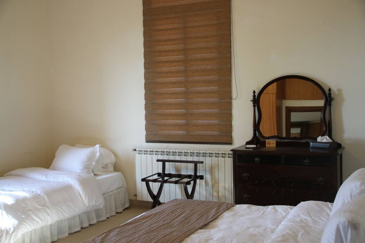 Ein Schlafzimmer mit zwei Einzelbetten, einer Holzkommode, einem Spiegel und einem Fenster mit Jalousien.