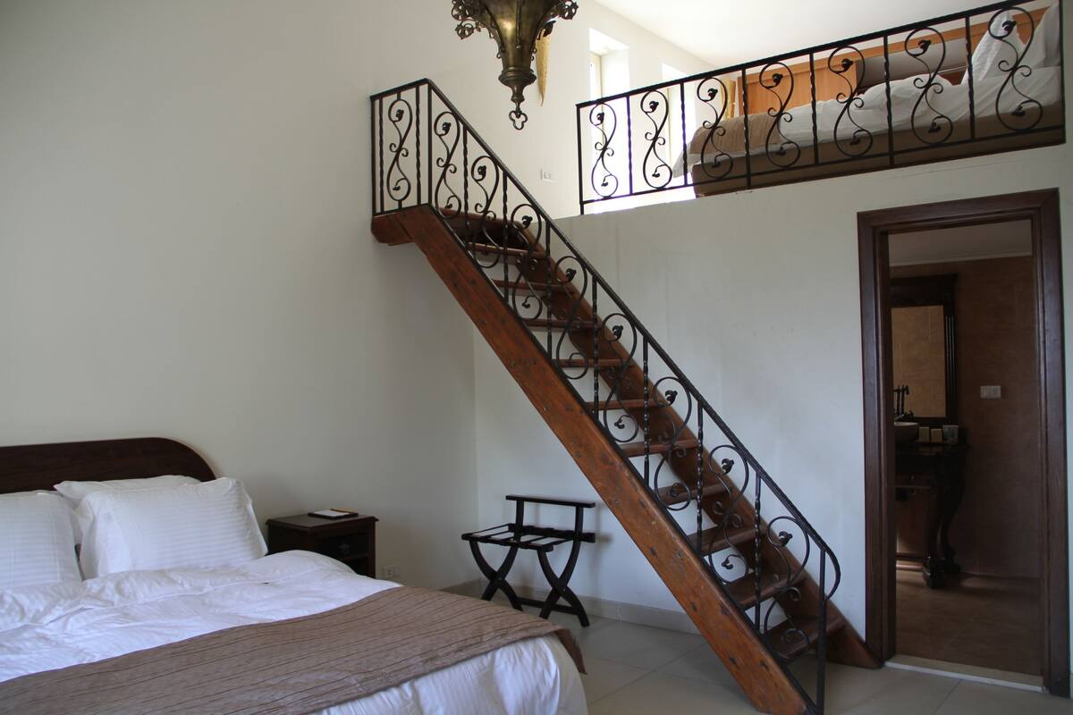 Ein Schlafzimmer mit einem hölzernen Bett, einem Nachttisch, einem kleinen Tisch und einer Treppe mit schmiedeeisernem Geländer.