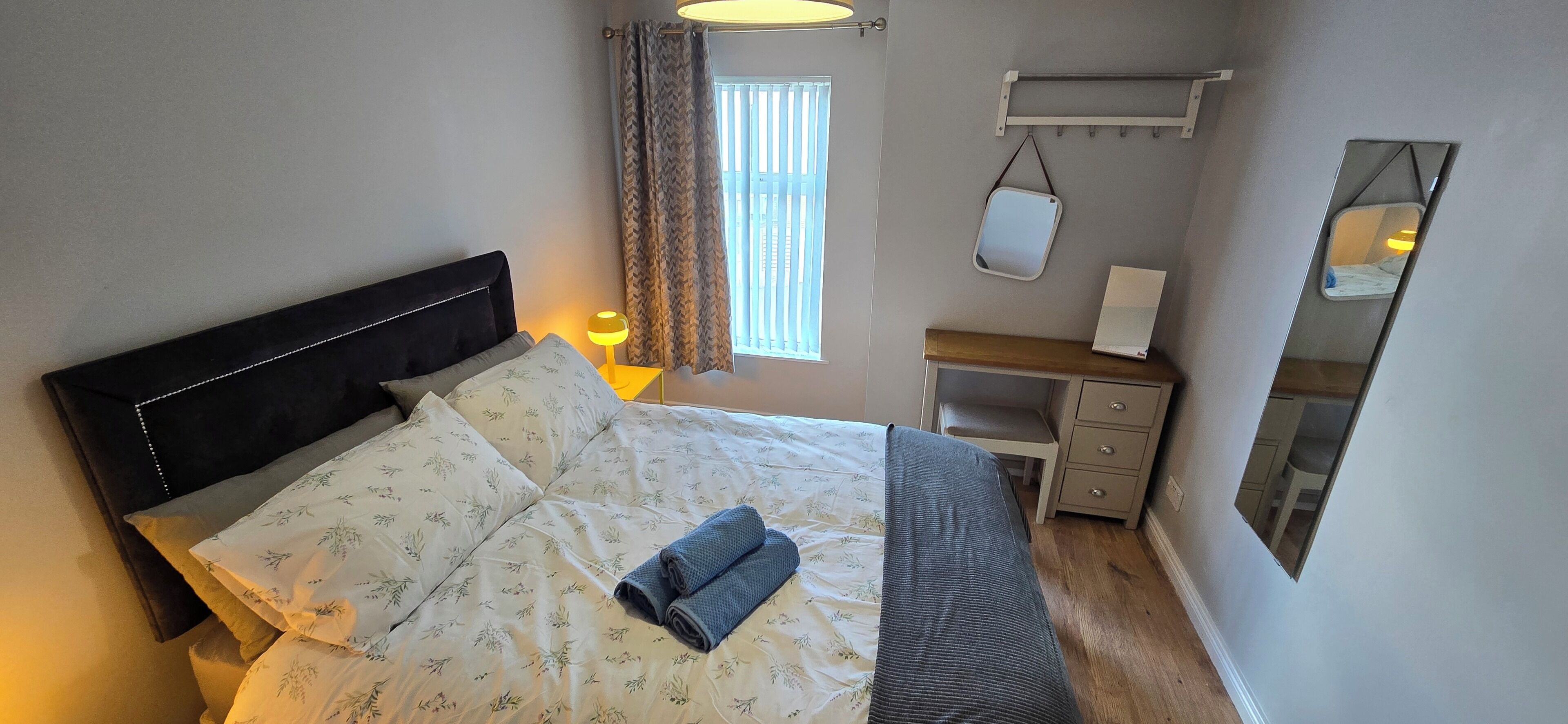 A neatly made bed with floral bedding, a bedside lamp, a small desk with a chair, and a mirror on the wall.