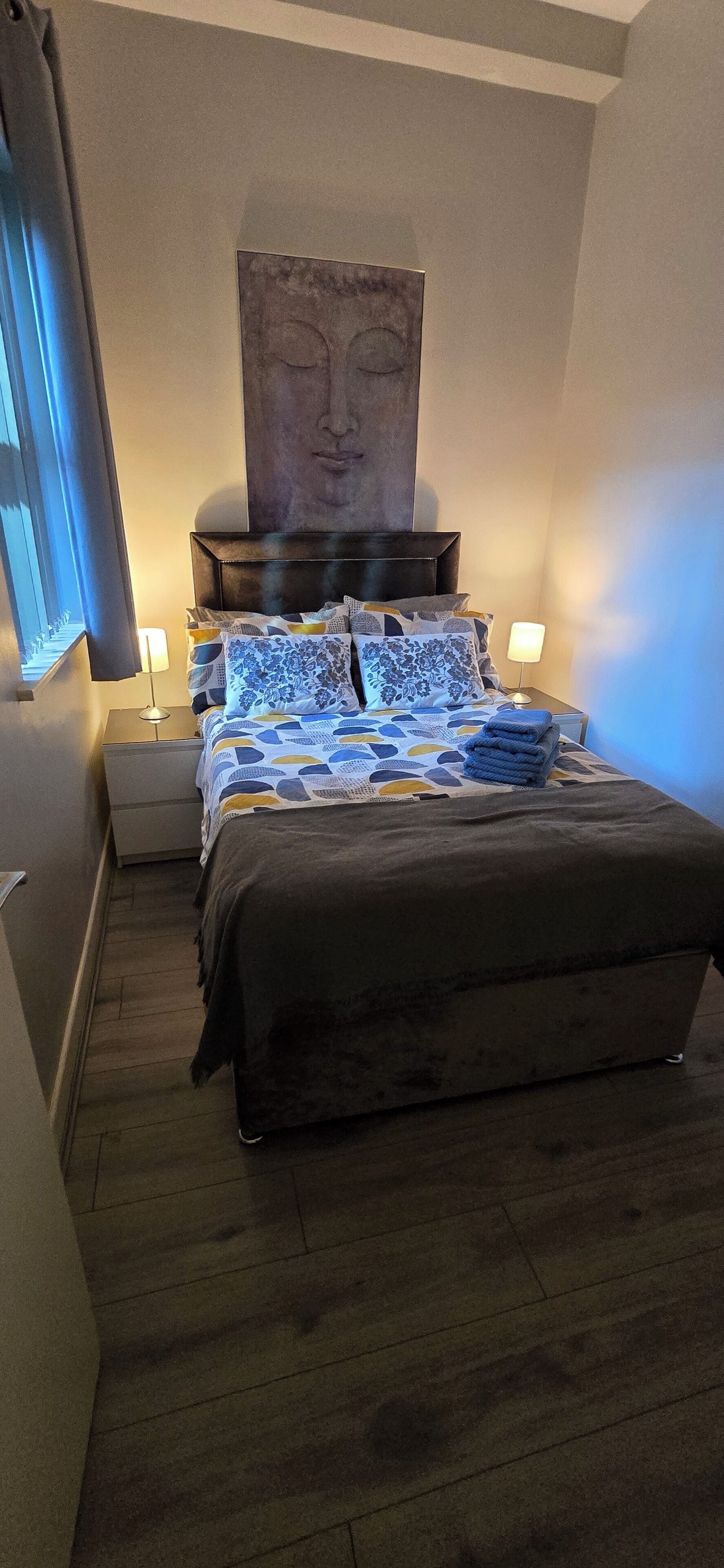 Ett hotellrum med en säng, en sänggavel, ett konstverk som hänger på väggen, sänglampor och ett fönster med gardiner.