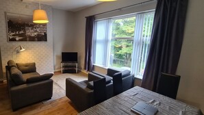 A modern living room with a sofa, armchairs, a dining table, a TV, and a large window with curtains. - Castleton Gardens Apartments Belfast (Belfast)