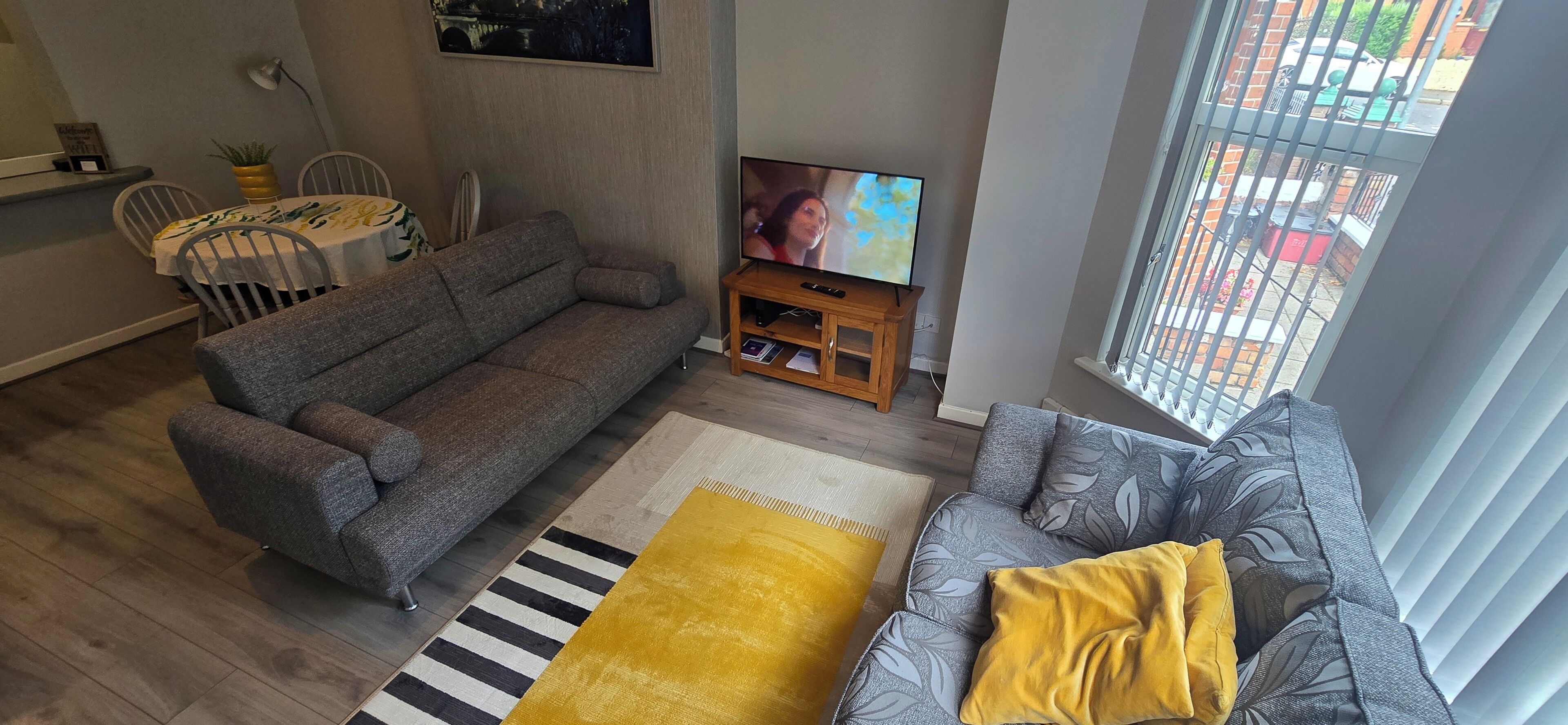 A living room with a grey sofa, a yellow ottoman, a striped rug, and a TV on a wooden stand.