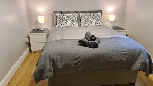 A neatly made bed with a grey blanket and two pillows, two bedside lamps, a small bedside table with a lamp, and a power outlet on the wall. - Castleton Gardens Apartments Belfast (Belfast)