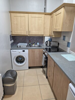 A compact kitchen with wooden cabinets, a washing machine, a coffee maker, and a toaster. - Castleton Gardens Apartments Belfast (Belfast)