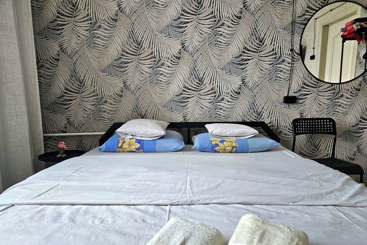 A bed with white bedding, two pillows with floral patterns, and a nightstand with a small table and a flower.