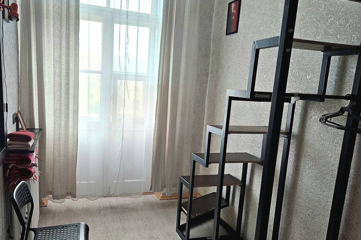 A room with a wooden floor, a black metal shelf unit, a black chair, and a window with white curtains.