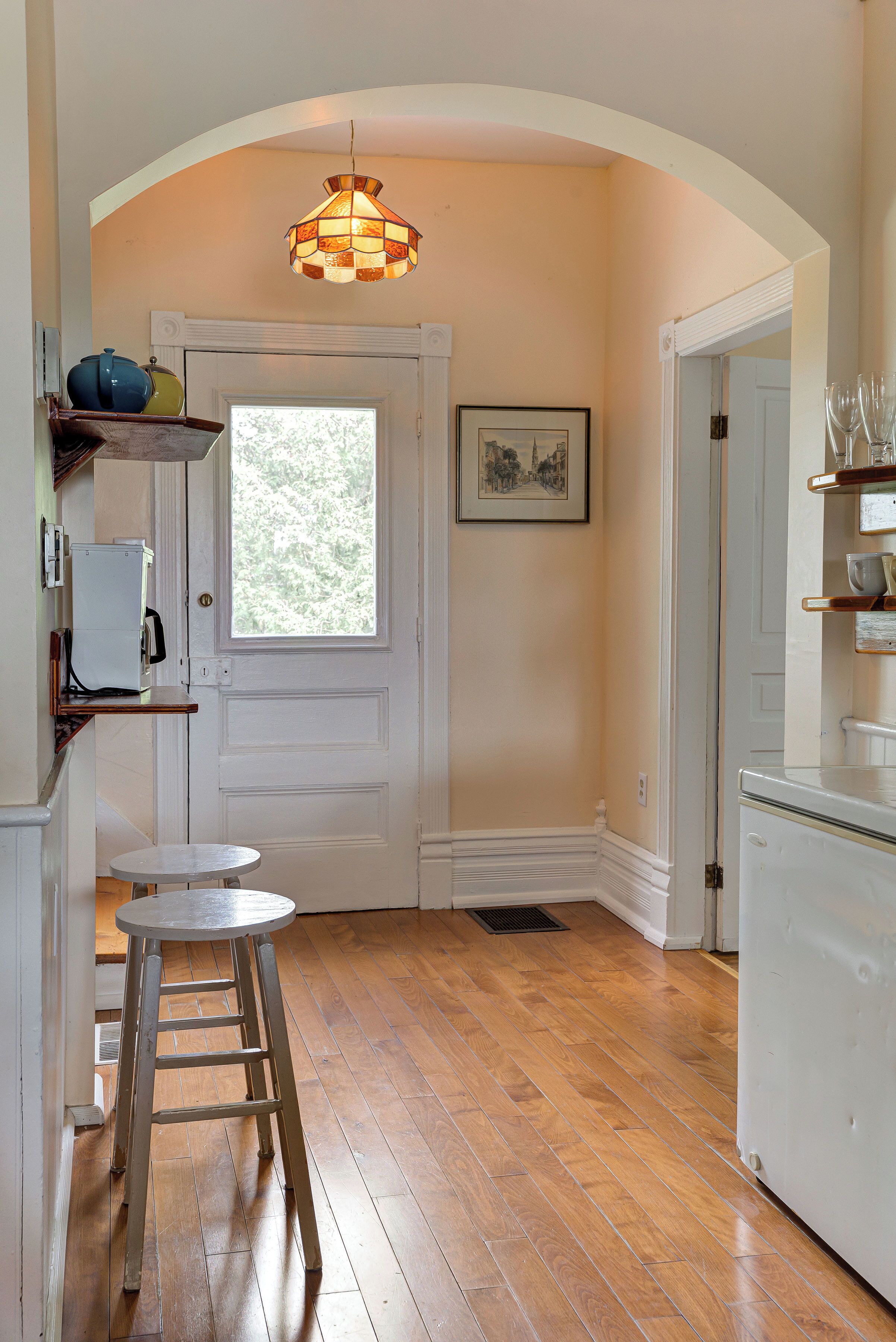 A kitchen with wooden flooring, a white door, a hanging light fixture, and a window.