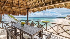 A wooden table and chairs under a thatched roof, overlooking a beach with clear turquoise water and a sandy shore.