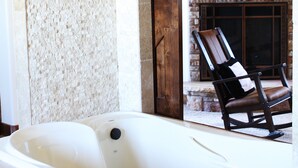 A stone-walled bathroom with a bathtub, a wooden door, and a fireplace with a chair in the background.