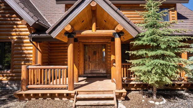 A log cabin with a wooden porch, a front door, and a small tree in front.