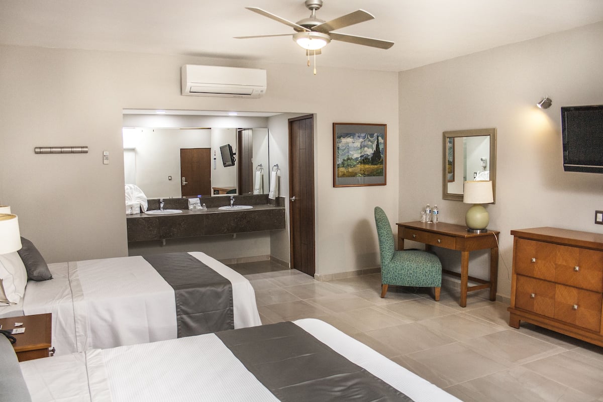 Une chambre d’hôtel avec deux lits, un bureau, une chaise, une commode, un ventilateur de plafond, un grand miroir et un téléviseur.