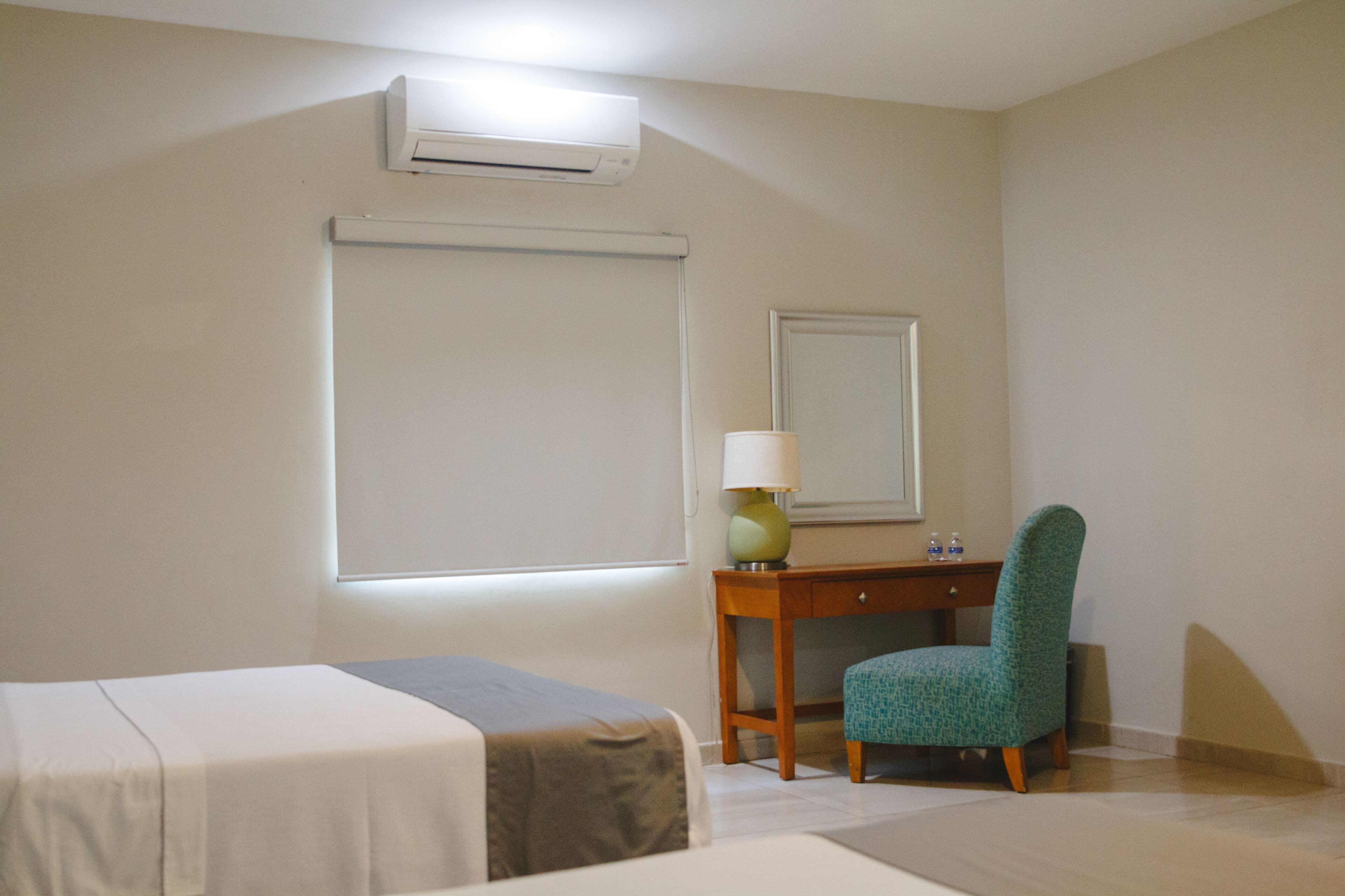 A hotel room with a bed, a desk with a chair, a lamp, and an air conditioning unit.