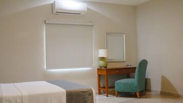 A hotel room with a bed, a desk with a chair, a lamp, and an air conditioning unit.