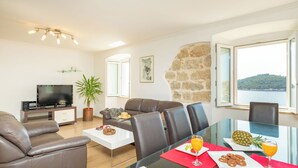 A modern living room with a sofa, dining table, and a TV. There is a view of the sea through large windows. - Hedera Estate, Hedera A19 (Dubrovnik)