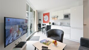 Una sala moderna con televisor de pantalla plana, comedor con mesa y sillas, cocina con gabinetes blancos y electrodomésticos de acero inoxidable, y un ventanal con cortinas.