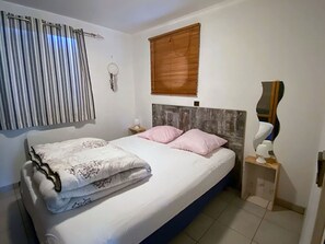 A bedroom with a bed, a nightstand, a mirror, a window with blinds, and a wall-mounted light fixture. - House, garden in Vieux Boucau (Vieux-Boucau-Les-Bains)
