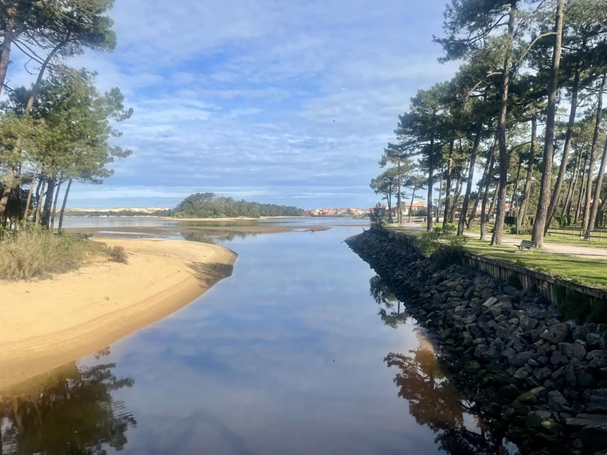 A calm river with a sandy bank and a stone wall on one side, surrounded by trees and a clear sky.