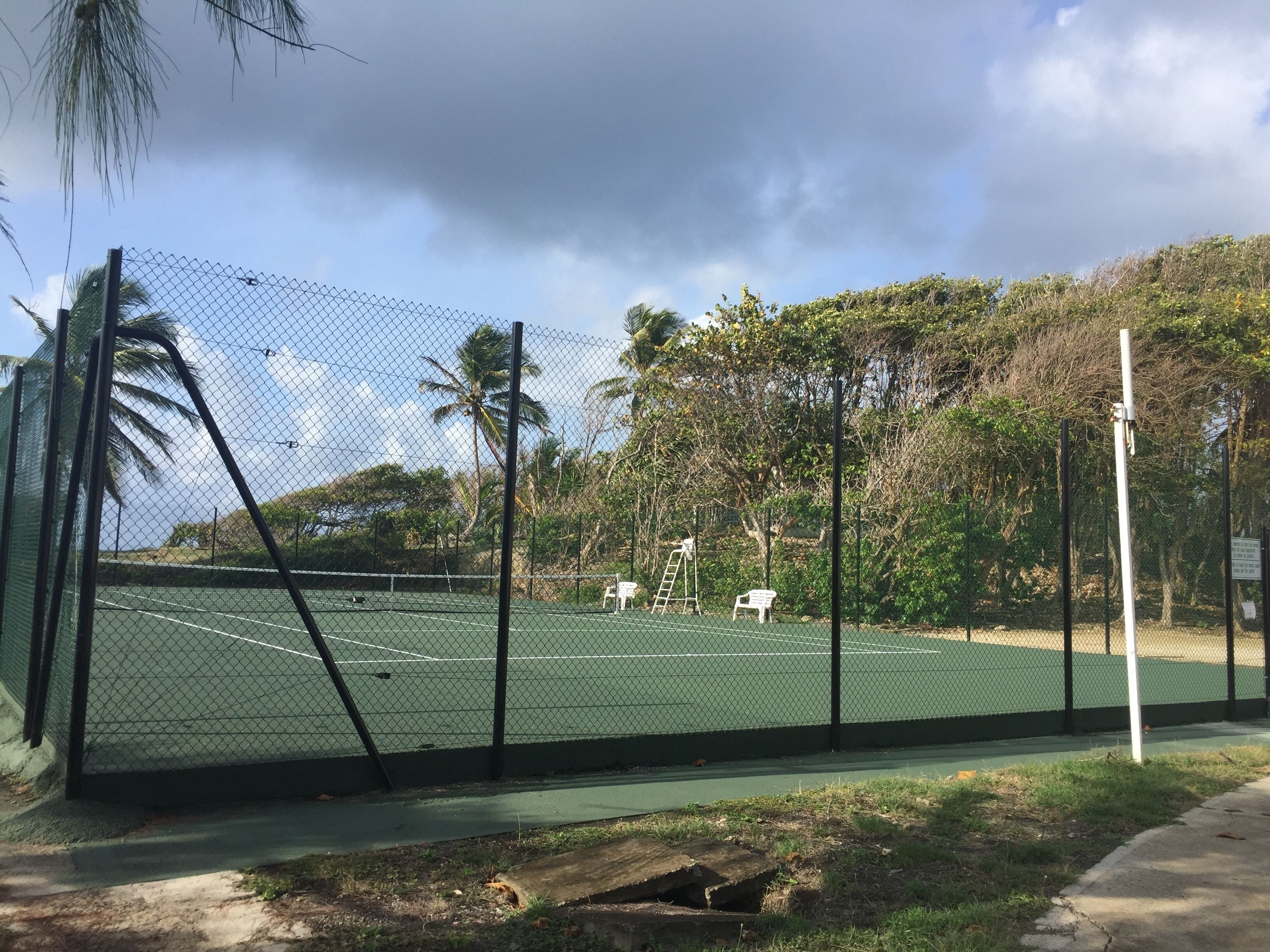 A tennis court surrounded by a chain-link fence, with a ladder and two white chairs nearby.