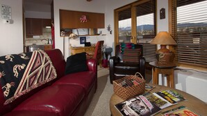 Sala de estar com sofá vermelho, mesa de centro de madeira com revistas, poltrona de couro e vista através de amplas janelas.