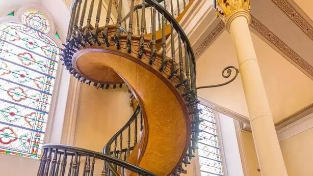 Uma escadaria em espiral com grades elaboradas e um corrimão de madeira, conduzindo a um andar superior em um edifício com janelas de vitrais e molduras decorativas.