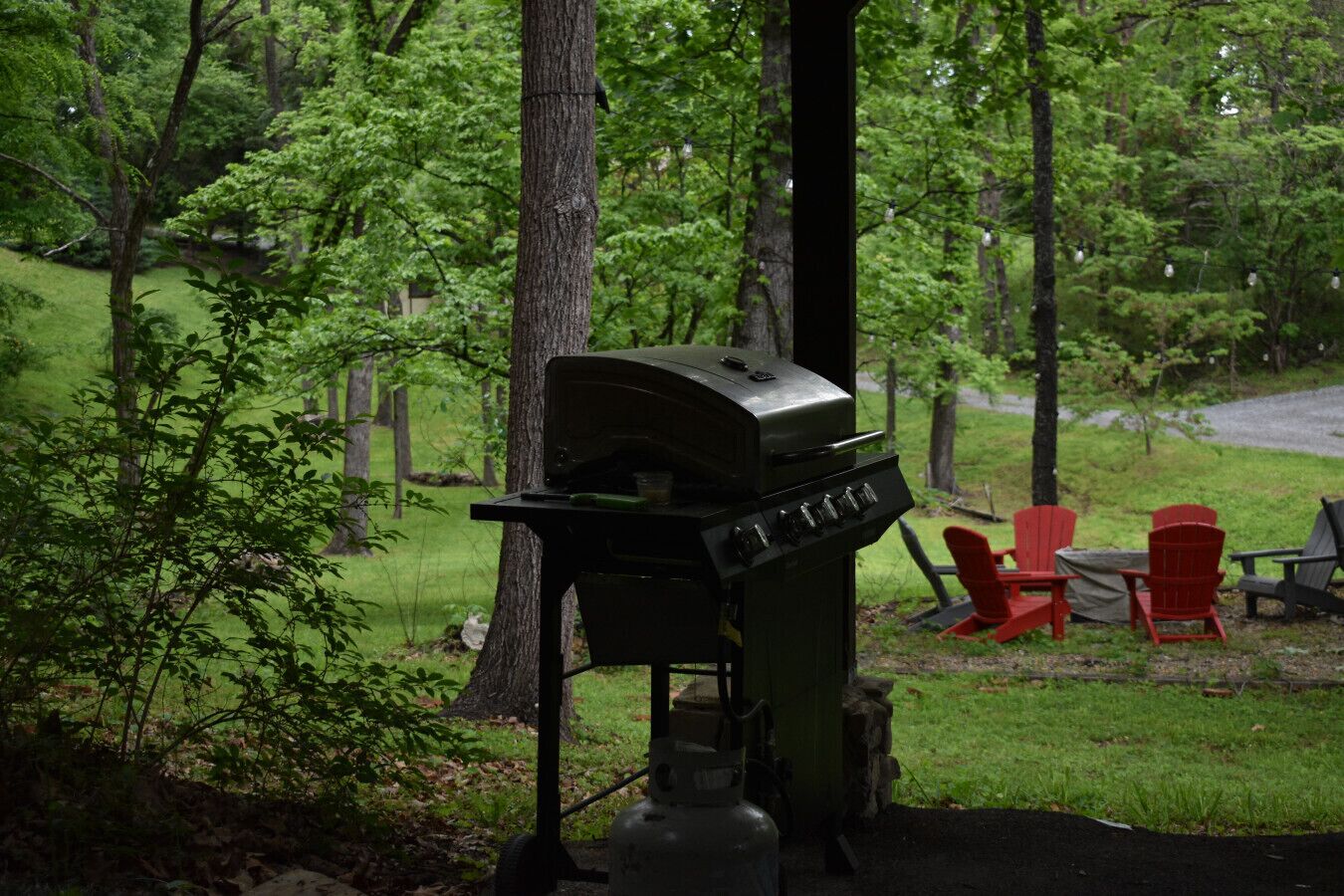 A grill in a wooded area with red chairs and a table.