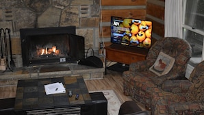 Ein gemütliches Wohnzimmer mit einem Kamin aus Stein, einem Fernseher und einem gemusterten Sessel.