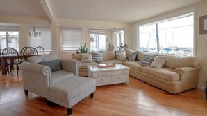 A spacious living room with a high ceiling, a dining area with a table and chairs, and large windows offering a view of the outside. - 6 Beds, 4 Baths w Elevator LBI - Now Booking 2027 limited 2026 dates still avail (Beach Haven)